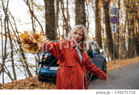 Bunch of fallen leaves, fun. Beautiful blonde woman is with her blue electric car on the road of forest Bunch of fallen leaves, fun. Beautiful blonde woman is with her blue electric car on the road of forest 139275710