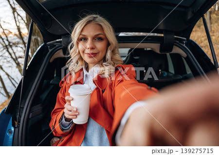 Selfie time. Positive facial expression. Beautiful blonde woman is with her blue electric car on the road of forest Selfie time. Positive facial expression. Beautiful blonde woman is with her blue electric car on the road of forest 139275715