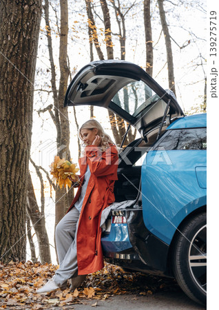 Bunch of fallen leaves, fun. Beautiful blonde woman is with her blue electric car on the road of forest Bunch of fallen leaves, fun. Beautiful blonde woman is with her blue electric car on the road of forest 139275719