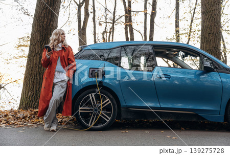 Automobile is charging. Beautiful blonde woman is with her blue electric car on the road of forest Automobile is charging. Beautiful blonde woman is with her blue electric car on the road of forest 139275728