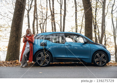 Automobile is charging. Beautiful blonde woman is with her blue electric car on the road of forest Automobile is charging. Beautiful blonde woman is with her blue electric car on the road of forest 139275729