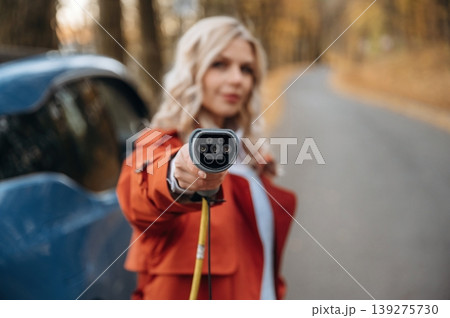 Holding the charging cable. Beautiful blonde woman is with her blue electric car on the road of forest Holding the charging cable. Beautiful blonde woman is with her blue electric car on the road of forest 139275730