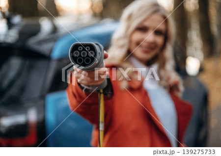 Holding the charging cable. Beautiful blonde woman is with her blue electric car on the road of forest Holding the charging cable. Beautiful blonde woman is with her blue electric car on the road of forest 139275733