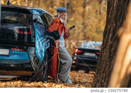 Black smartphone in hands, standing and waiting for charging to complete. Beautiful blonde woman is with her blue electric car on the road of forest Black smartphone in hands, standing and waiting for charging to complete. Beautiful blonde woman is with her blue electric car on the road of forest 139275740