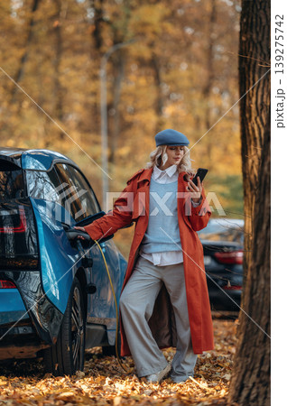 Rural non urban scene. Waiting for auto to charge. Beautiful blonde woman is with her blue electric car on the road of forest Rural non urban scene. Waiting for auto to charge. Beautiful blonde woman is with her blue electric car on the road of forest 139275742