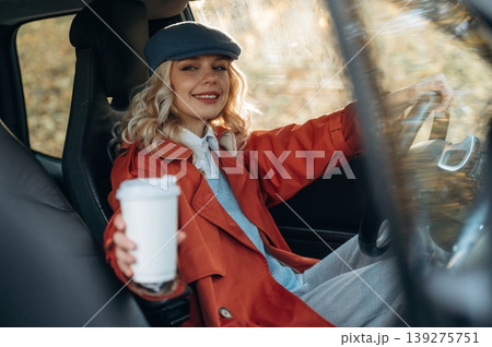 Hot drink in white cup. Happy blonde woman is sitting her electric car Hot drink in white cup. Happy blonde woman is sitting her electric car 139275751