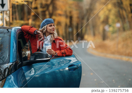 In blue beret. Beautiful blonde woman is with her blue electric car on the road of forest In blue beret. Beautiful blonde woman is with her blue electric car on the road of forest 139275756