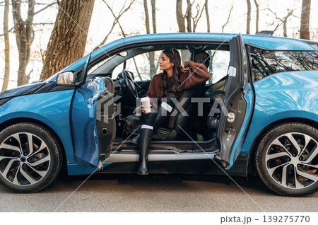 On the road, door opened, parked. Beautiful woman is sitting in her electric car On the road, door opened, parked. Beautiful woman is sitting in her electric car 139275770