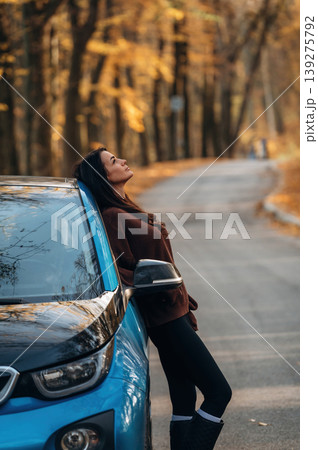 In brown sweater and black pants. Woman is outdoors near her electric blue car at daytime, rural scene In brown sweater and black pants. Woman is outdoors near her electric blue car at daytime, rural scene 139275792