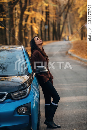 In brown sweater and black pants. Woman is outdoors near her electric blue car at daytime, rural scene In brown sweater and black pants. Woman is outdoors near her electric blue car at daytime, rural scene 139275793