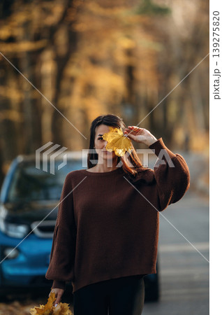 Leaves are in hands. Woman is outdoors near her electric blue car at daytime, rural scene 139275820