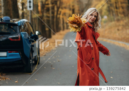 Bunch of fallen leaves, fun. Beautiful blonde woman is with her blue electric car on the road of forest Bunch of fallen leaves, fun. Beautiful blonde woman is with her blue electric car on the road of forest 139275842