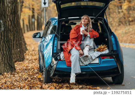 Cup of drink, sitting in the trunk. Beautiful blonde woman is with her blue electric car on the road of forest Cup of drink, sitting in the trunk. Beautiful blonde woman is with her blue electric car on the road of forest 139275851