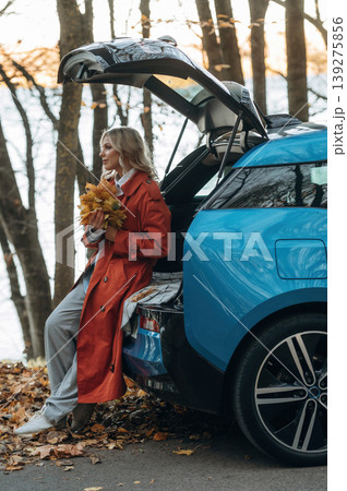 Having fun with fallen leaves. Beautiful blonde woman is with her blue electric car on the road of forest Having fun with fallen leaves. Beautiful blonde woman is with her blue electric car on the road of forest 139275856