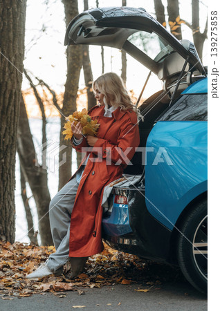 The fallen leaves are in hands. Beautiful blonde woman is with her blue electric car on the road of forest 139275858