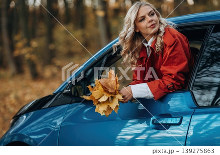 The fallen leaves are in hands. Beautiful blonde woman is with her blue electric car on the road of forest The fallen leaves are in hands. Beautiful blonde woman is with her blue electric car on the road of forest 139275863