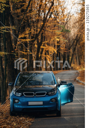 Front view of blue electric car with door opened, parked on the sideroad, with orange colored autumn trees at background Front view of blue electric car with door opened, parked on the sideroad, with orange colored autumn trees at background 139275865