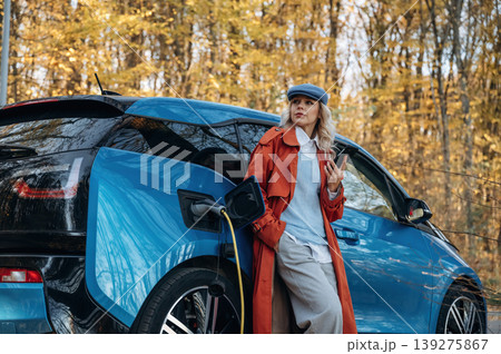 Waiting for the charging to complete. Beautiful blonde woman is with her blue electric car on the road of forest 139275867