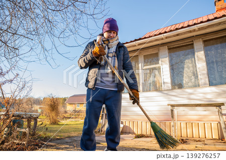 Man sweeping the backyard with a broom. Yard work and outdoor house maintenance in autumn. Home improvement and spring cleaning concept. Caucasian guy tidying the rural patio area. 139276257