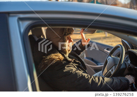 Man in winter hat adjusting rearview mirror inside car. Driver preparing for travel during cold weather. Safety check before departure. Transportation and vehicle maintenance concept. 139276258