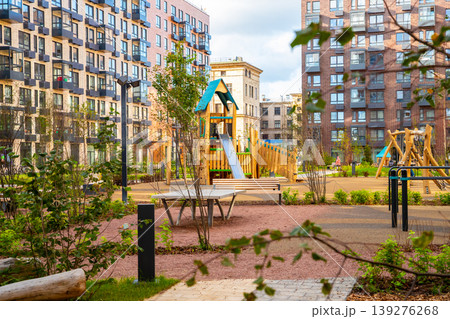 Modern urban playground courtyard between residential brick apartment buildings. Neighborhood outdoor recreational space with park bench, ping pong table and wooden climbing frame structure. Modern urban playground courtyard between residential brick apartment buildings. Neighborhood outdoor recreational space with park bench, ping pong table and wooden climbing frame structure. 139276268
