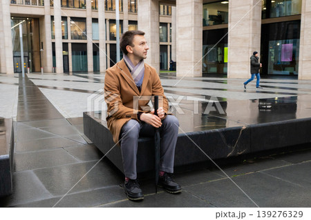 Office worker sitting on stone bench outdoor holding umbrella while waiting for job interview. Man in coat relaxing during break near financial center. Professional lifestyle concept. Office worker sitting on stone bench outdoor holding umbrella while waiting for job interview. Man in coat relaxing during break near financial center. Professional lifestyle concept. 139276329