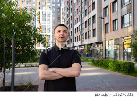Young man entrepreneur standing with arms crossed in front of modern apartment building. Successful businessman looking away. City lifestyle, apartment investment and urban development. Young man entrepreneur standing with arms crossed in front of modern apartment building. Successful businessman looking away. City lifestyle, apartment investment and urban development. 139276332
