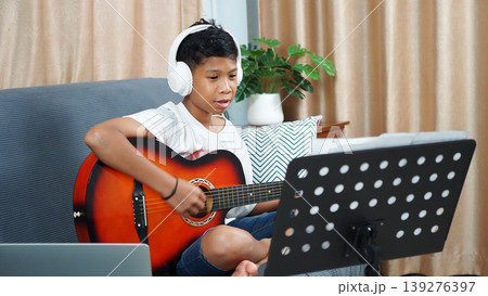 Child learning musical instrument via video call on laptop,Digital education concept with kid playing acoustic guitar,Asian boy taking online guitar lesson with headphones. 139276397
