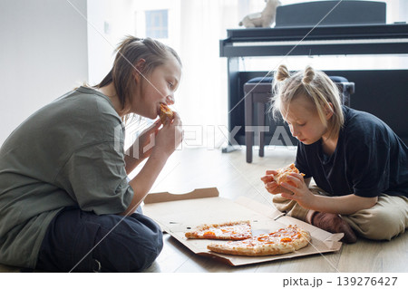 Adorable young children sitting on floor in living room and eating pizza at home Adorable young children sitting on floor in living room and eating pizza at home 139276427