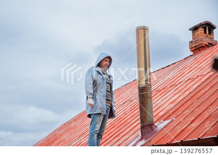 Chimney sweep standing on red metal roof. Man inspecting chimney pipe for professional maintenance and cleaning service. Repair of heating system and furnace chimney on house exterior. Chimney sweep standing on red metal roof. Man inspecting chimney pipe for professional maintenance and cleaning service. Repair of heating system and furnace chimney on house exterior. 139276557