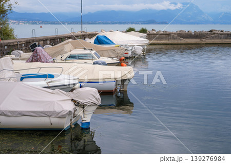 Row of small boats covered with tarps moored in calm harbor, with scenic lake and mountains in background, peaceful waterfront landscape scene. Row of small boats covered with tarps moored in calm harbor, with scenic lake and mountains in background, peaceful waterfront landscape scene. 139276984