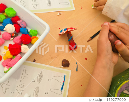A child molds colorful clay into a mushroom shape at a classroom desk. Concepts of creativity, tactile learning, and child development through play. 139277000