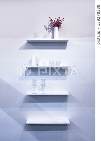 White wooden shelves mounted on wall with decorative flower vase and clear glass tableware. Minimalist interior design concept. Modern home decor for living room organization storage. White wooden shelves mounted on wall with decorative flower vase and clear glass tableware. Minimalist interior design concept. Modern home decor for living room organization storage. 139278388