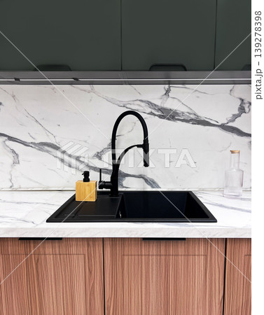 Modern kitchen sink with black faucet and soap dispenser on white marble countertop. Contemporary home interior design element with wood cabinets and backsplash. Minimalist kitchen style. Modern kitchen sink with black faucet and soap dispenser on white marble countertop. Contemporary home interior design element with wood cabinets and backsplash. Minimalist kitchen style. 139278398