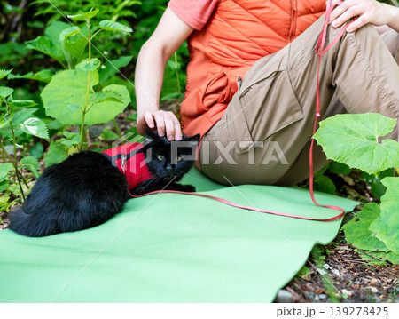 Woman sitting on green yoga mat in forest petting black cat in red harness. Owner relaxing with pet outdoors on summer day. Leisure activity, animal care and companionship concept. 139278425
