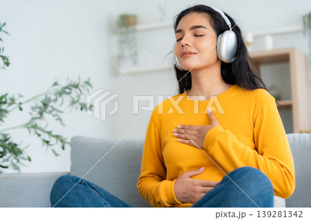 Happy young Asian woman practicing yoga and meditation at home sitting on couch in living room Happy young Asian woman practicing yoga and meditation at home sitting on couch in living room 139281342