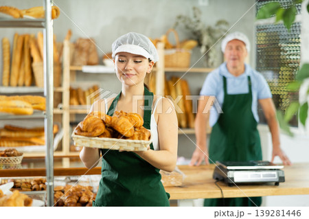Portrait of young happy girl seller in apron working in bakehouse, holding tray with fresh croissants on bakery shop Portrait of young happy girl seller in apron working in bakehouse, holding tray with fresh croissants on bakery shop 139281446