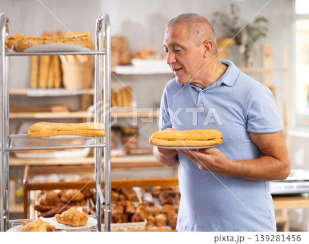 Senior male customer stands with croissant in hands near window of bakery. Senior male customer stands with croissant in hands near window of bakery. 139281456
