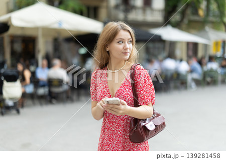Young woman in summer dress standing on street with phone in hands Young woman in summer dress standing on street with phone in hands 139281458