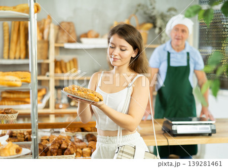 Positive beautiful girl buyer choosing and holding bocadillo in hands, elderly man seller standing in background in bakery shop Positive beautiful girl buyer choosing and holding bocadillo in hands, elderly man seller standing in background in bakery shop 139281461
