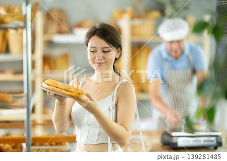 Happy girl buyer standing small trading room of bakery with bocadillo in hands, elderly male baker working in background Happy girl buyer standing small trading room of bakery with bocadillo in hands, elderly male baker working in background 139281485
