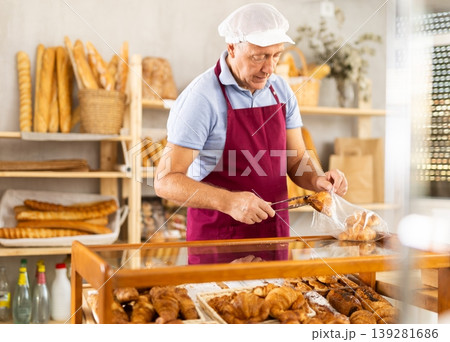 Senior man seller puts croissant in plastic disposable bag for buyer, packs goods. Senior man seller puts croissant in plastic disposable bag for buyer, packs goods. 139281686