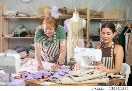 Elderly dressmaker making marks on fabric next to young seamstress 139281852