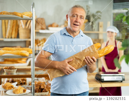 European aged male buyer stands proudly in front of fresh bread holding freshly baked baguettes in bakery store 139282531