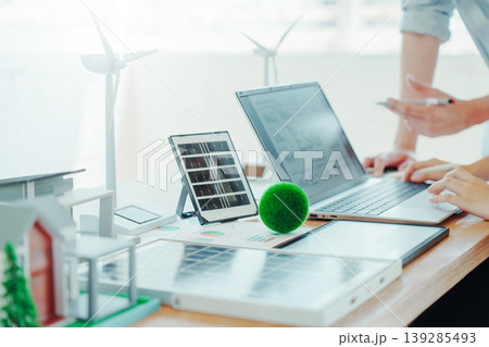 Renewable energy workspace with laptop, solar panel model, wind turbines and eco house mockup on desk, people analyzing data and planning sustainable power project in bright office. 139285493