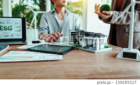 Business meeting on sustainable real estate: ESG dashboard on laptop, model eco house on desk, wind turbines and green concept props in modern office. Business meeting on sustainable real estate: ESG dashboard on laptop, model eco house on desk, wind turbines and green concept props in modern office. 139285527
