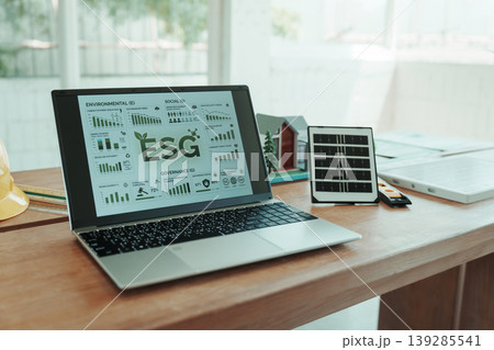 Laptop on office desk showing esg analytics dashboard with charts; tablet with grid display beside it in bright modern workspace for sustainability, corporate reporting and governance concept. 139285541