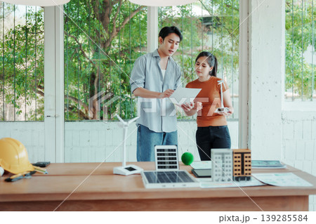 Two young professionals review plans in a bright office with renewable energy and building models on desk, green outdoor view through large windows. Two young professionals review plans in a bright office with renewable energy and building models on desk, green outdoor view through large windows. 139285584