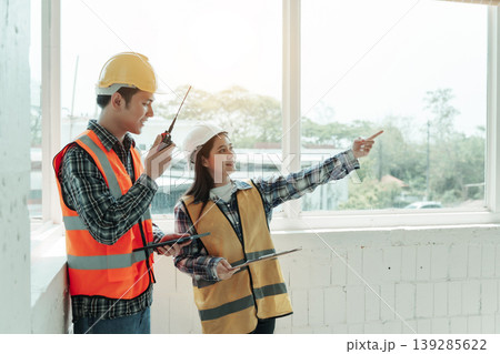 Two construction workers in hard hats and safety vests coordinate indoors by large window, the other points, suggesting site inspection and planning. 139285622