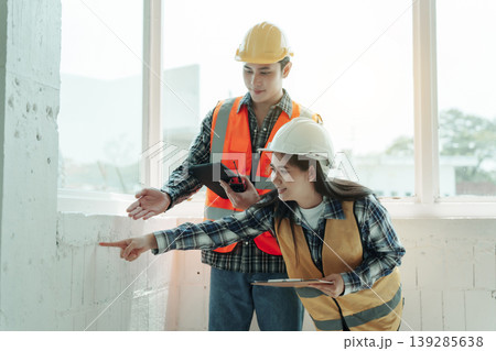 Two construction workers in hard hats and safety vests inspecting an interior wall at a building site, using a tablet and clipboard in bright natural light. Two construction workers in hard hats and safety vests inspecting an interior wall at a building site, using a tablet and clipboard in bright natural light. 139285638